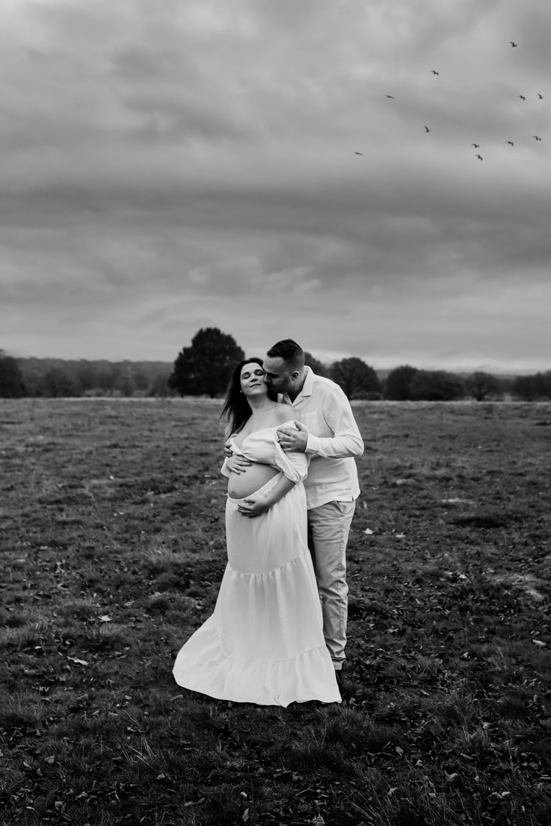 London Family Photographer, a man kisses his partner, she is smiling and holds her pregnant belly at the park