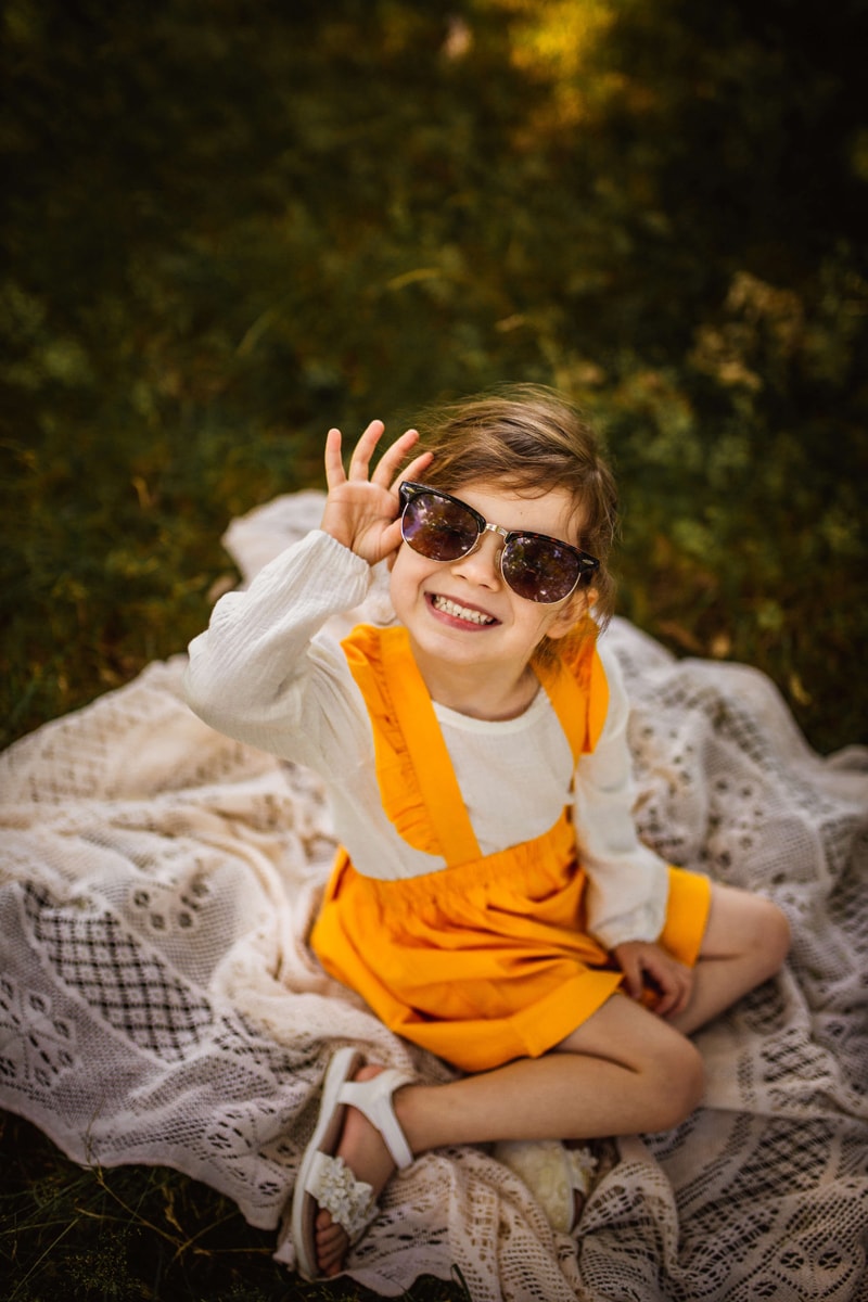 London Family Photographer, a young girl smiles as she holds onto a large pair of sunglasses on her face, she sits on a blanket