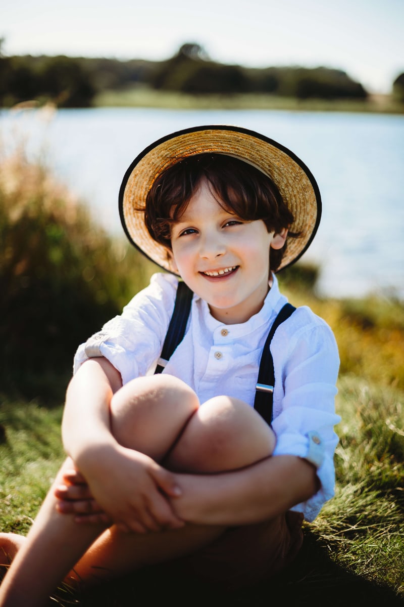 London Family Photographer, a young boy sits in the grass with a straw hat near a lake