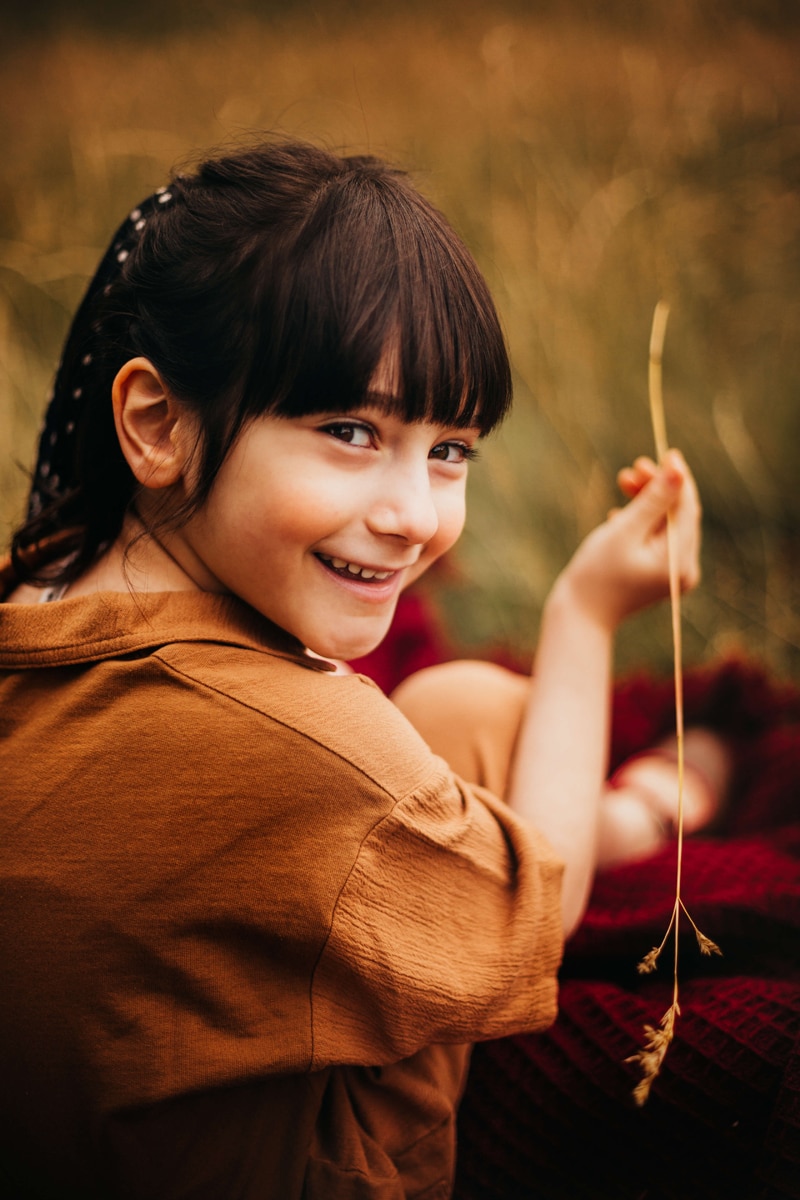 London Family Photographer, a young smiling girl holds up a piece of straw she has plucked