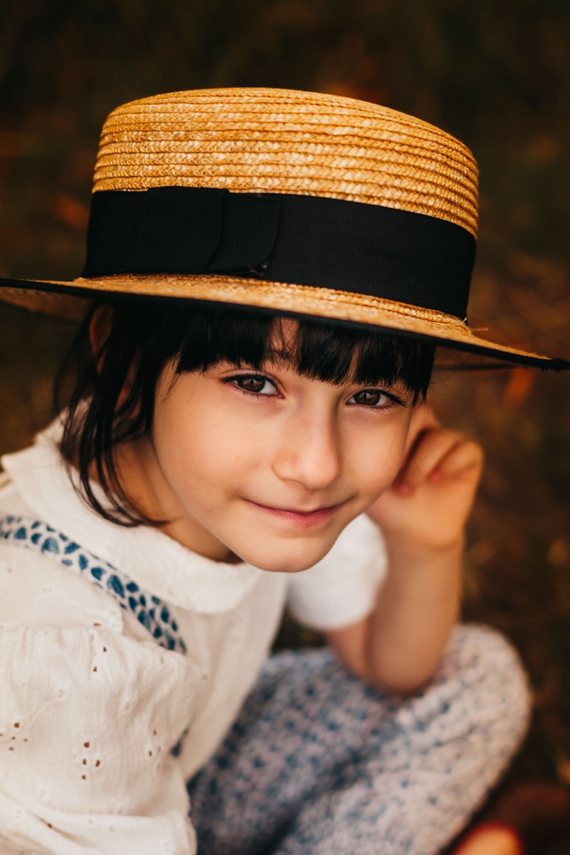 London Family Photographer, a young child sits pondering, smiling, and wearing a straw hat