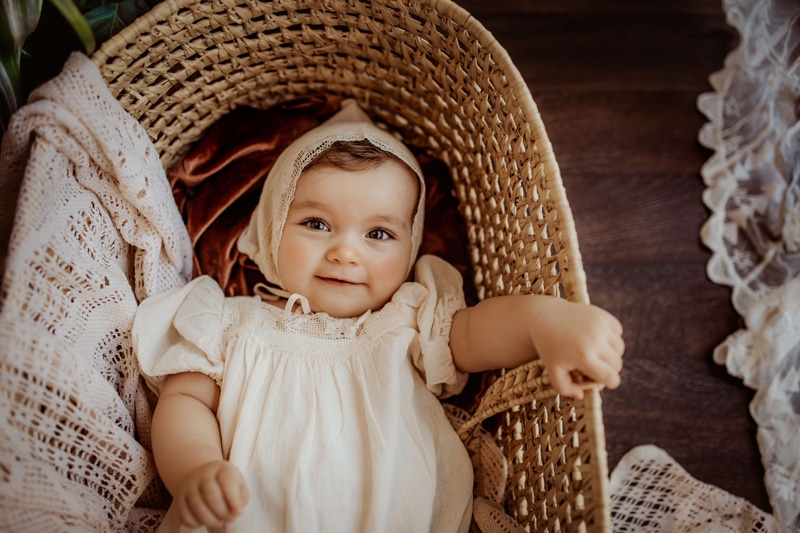 London Family Photographer, a baby lays smiling in a basket, her hand grips the wicker edge