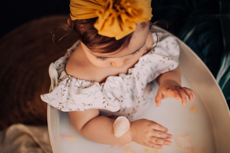 London Family Photographer, a young girl dressed in flowery attire and a large bow, sits in a milk bath with flower pedals on her hands