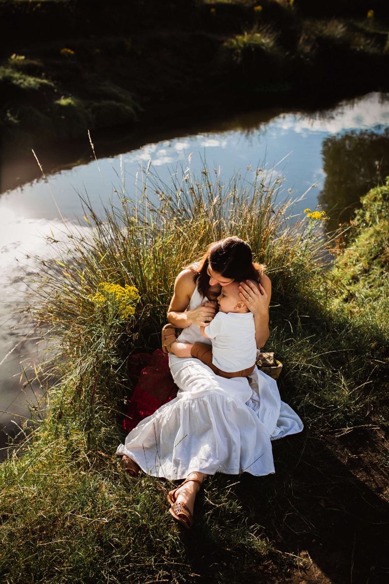 London Family Photographer, a young mother in a white dress holds onto her newborn baby sun near a stream
