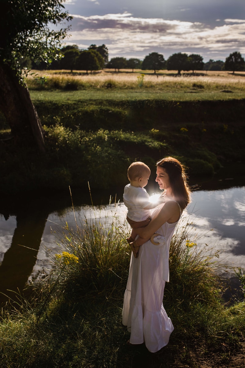 London Family Photographer, a woman holds her baby near a quiet stream, the clouds reflect in the water