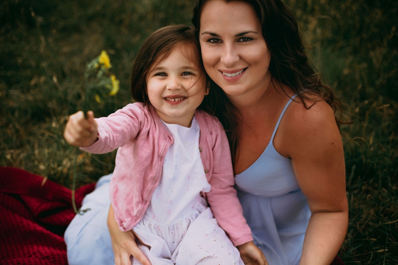 London Family Photographer, a mom smiles holding onto her young daughter, the daughter holds freshly-picked flowers